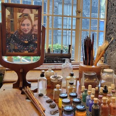 Frida Kahlo's rectangular mirror is on a table surrounded by glass bottles containing bright paint pigment. There is a pot containing her paintbrushes. The reflection in the mirror is Red and she is holding her phone taking a photo. In the background