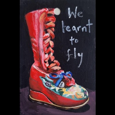 We Learnt to Fly is written in white text to the top right of this painting of a red boot. The boot belonged to Frida Kahlo and has embroidered flowers and shapes on the front of the boot and a bell is attached with a sky blue ribbon